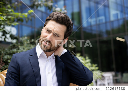 Close-up portrait of a young businessman standing in a suit near an office building and holding his neck, tired after a working day, feeling pain, massaging his hand. Close-up portrait of a young businessman standing in a suit near an office building and holding his neck, tired after a working day, feeling pain, massaging his hand. 112617188