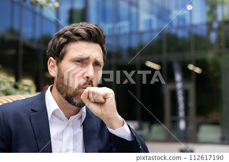 Close-up photo of a young sick man in a business suit who is standing outside an office building and coughing, covering his mouth with his hand. Close-up photo of a young sick man in a business suit who is standing outside an office building and coughing, covering his mouth with his hand. 112617190