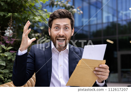 Close-up portrait of a happy and shocked young man in a business suit sitting near an office building outside, holding a torn envelope with a letter in his hands and shouting at the camera with joy. 112617207