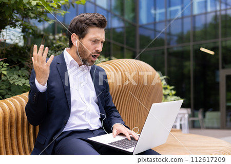 Dissatisfied young pencil man in a suit and headphones sitting outside on a bench and talking angrily on a video call on a laptop, throwing up his hands in frustration. Dissatisfied young pencil man in a suit and headphones sitting outside on a bench and talking angrily on a video call on a laptop, throwing up his hands in frustration. 112617209
