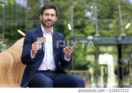 Portrait of a smiling young businessman and banker sitting on a bench outside an office building in a suit, holding a telephone and showing a credit card to the camera. 112617212