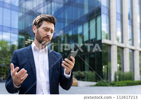 Close-up photo of a disappointed young businessman looking at the phone screen and upset by the received news, standing outside the office and spreading his hands. 112617213