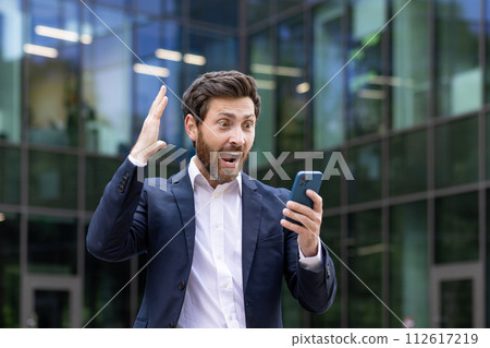 Photo of a shocked young man in a business suit standing on the street and looking at the phone screen in surprise, shouting with happiness, raising his hand up. 112617219