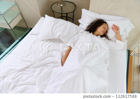 View from above of a woman sleeping in the bed, covered with white soft blanket. 112617588