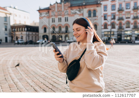 Closeup portrait of a lovely young girl listening to music through wireless earphones on urban background. Music lover enjoying music. Woman record voice by mobile cell phone dictaphone Closeup portrait of a lovely young girl listening to music through wireless earphones on urban background. Music lover enjoying music. Woman record voice by mobile cell phone dictaphone 112617707