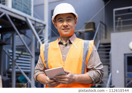 A professional industrial worker wearing a high-visibility vest holds a tablet while standing at an industrial site. A professional industrial worker wearing a high-visibility vest holds a tablet while standing at an industrial site. 112618544