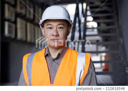 Confident Asian engineer in safety vest and hard hat standing in an industrial construction setting, portraying professionalism and expertise. 112618546