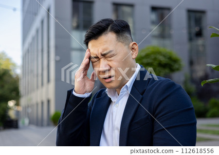 Worried Asian businessman in a blue suit feeling a headache, standing outside an office building with a pained expression. 112618556
