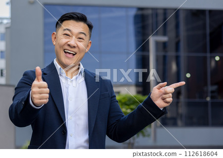 Cheerful Asian businessman in suit giving a thumbs up and pointing to the side, expressing approval and direction. Cheerful Asian businessman in suit giving a thumbs up and pointing to the side, expressing approval and direction. 112618604