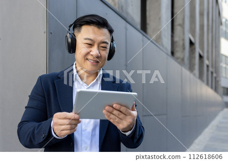 Professional male in a suit multitasking with a digital tablet and headphones outside a modern building. Professional male in a suit multitasking with a digital tablet and headphones outside a modern building. 112618606