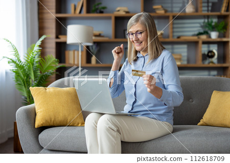 Joyful woman sitting on couch with laptop, raising fist while holding a credit card, feeling victorious after an online transaction. Joyful woman sitting on couch with laptop, raising fist while holding a credit card, feeling victorious after an online transaction. 112618709