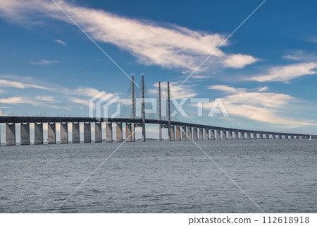 The Oresund Bridge, connecting Copenhagen, Denmark, with Malmo, Sweden, arches over calm waters, its high pylons and cables creating a striking silhouette against a clouddotted sky. 112618918
