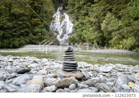 Stack of rocks, cairn, positioned on a pebble beach in front of big waterfall , New Zealand Stack of rocks, cairn, positioned on a pebble beach in front of big waterfall , New Zealand 112623073