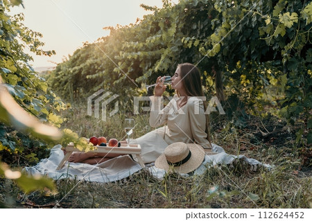 Woman picnic vineyard. Happy woman with a glass of wine at a picnic in the vineyard, wine tasting at sunset and open nature in the summer. Romantic dinner, fruit and wine. 112624452