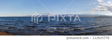 Blue calm water in Issyk-Kul lake with mountains on background at summer day - wide panorama 112625770