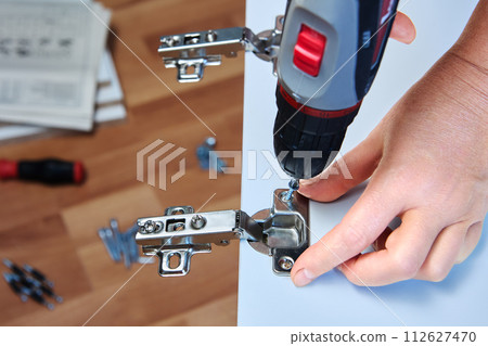 DIY installation adjustable door hinges on cabinet of flat pack, using power screwdriver. 112627470