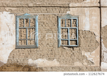 old house adobe air brick wall with two embedded wooden window frames and peeled off plaster leftovers - flat texture and full frame background 112627674