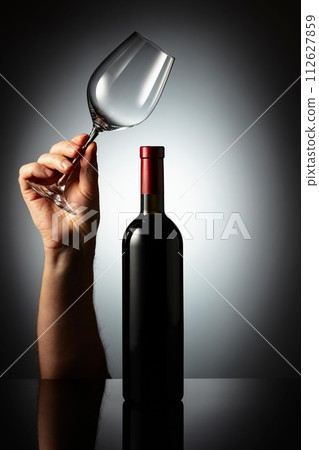 Unopened bottle of red wine and hand with wine glass. 112627859