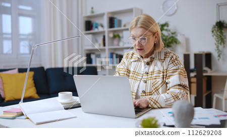 Serious mature woman focused on her work on laptop, remote financial analyst Serious mature woman focused on her work on laptop, remote financial analyst 112629208