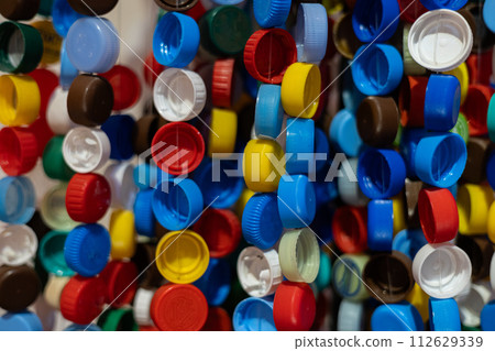 Multi-colored plastic caps hang in a row on a thread to form a beautiful backdrop. Waste plastic recycling. Composition of multi-colored plastic caps on a rope. Multi-colored plastic caps hang in a row on a thread to form a beautiful backdrop. Waste plastic recycling. Composition of multi-colored plastic caps on a rope. 112629339