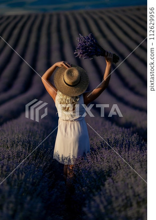Blonde woman poses in lavender field at sunset. Happy woman in white dress holds lavender bouquet. Aromatherapy concept, lavender oil, photo session in lavender 112629956