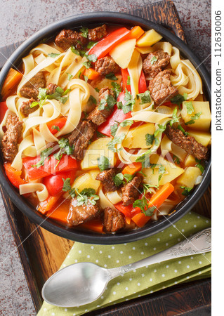 Laghman noodle soup from Uzbekistan made with beef chuck, tomato paste, vegetables and herbs closeup on the plate on the wooden board. Vertical top view Laghman noodle soup from Uzbekistan made with beef chuck, tomato paste, vegetables and herbs closeup on the plate on the wooden board. Vertical top view 112630006