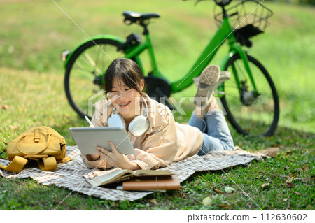 Smiling young woman lying on blanket in the public park and using digital tablet 112630602