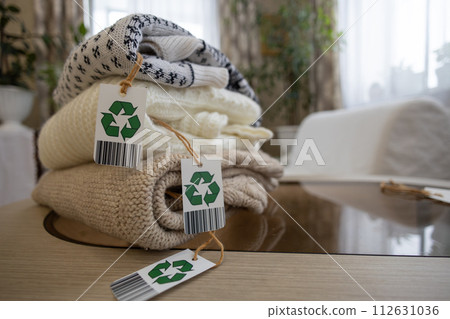 Tags with green recycling arrows on clothes folded on a table in the room. Using environmentally friendly clothing made from natural materials. Selection of old clothes for recycling. 112631036