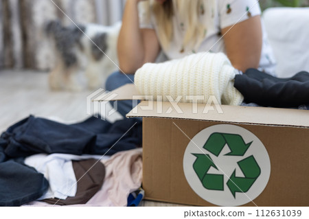 A box with a recycling sign with folded clothes stands on the floor. A woman sits in the background and goes through her wardrobe, sorting clothes. Selection of clothes for recycling. 112631039