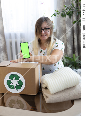 Vertical. A woman shows a green phone screen over a box of clothes for donation. Advertising for recycling, donation, clothing donation. Nice woman promoting clothing recycling. Vertical. A woman shows a green phone screen over a box of clothes for donation. Advertising for recycling, donation, clothing donation. Nice woman promoting clothing recycling. 112631045