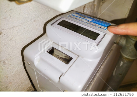 A household water meter in the entrance of a residential building, the finger points to the display showing how much has been consumed. Utility bills, regularity of expenses, family budget 112631796