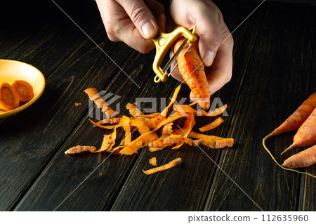 Chef peels raw carrots for a vegetable dish. Close-up of a chef's hands with a vegetable peeler on a black table Chef peels raw carrots for a vegetable dish. Close-up of a chef's hands with a vegetable peeler on a black table 112635960
