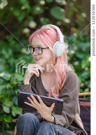 A cute pink-haired girl drawing on a digital tablet and wearing headphones in the garden, a Woman Doing Freelance Work in the Garden, and a woman with a digital tablet with artwork. 112636366