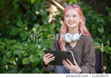 A cute pink-haired girl drawing on a digital tablet and wearing headphones in the garden, a Woman Doing Freelance Work in the Garden, and a woman with a digital tablet with artwork. A cute pink-haired girl drawing on a digital tablet and wearing headphones in the garden, a Woman Doing Freelance Work in the Garden, and a woman with a digital tablet with artwork. 112636374