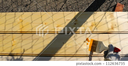 hand holds a wide brush with brown paint over a wooden space of planed boards on the street in sunny weather with shadows on a wooden surface, painting and protecting the wooden material 112636735
