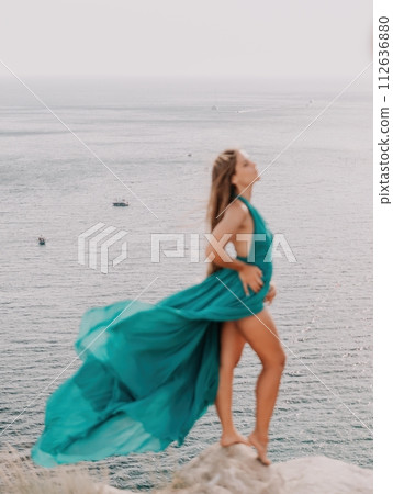 Woman travel portrait. Happy woman with long hair looking at camera and smiling. Close up portrait cute woman in a mint long dress posing on a volcanic rock high above the sea Woman travel portrait. Happy woman with long hair looking at camera and smiling. Close up portrait cute woman in a mint long dress posing on a volcanic rock high above the sea 112636880