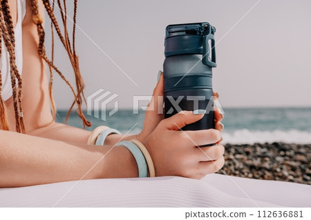 Woman sea yoga. Thermo mug in the hands of woman with braids dreadlocks in white leggings and tops doing stretching pilates on yoga mat near sea. Female fitness yoga routine concept. Healthy lifestyle Woman sea yoga. Thermo mug in the hands of woman with braids dreadlocks in white leggings and tops doing stretching pilates on yoga mat near sea. Female fitness yoga routine concept. Healthy lifestyle 112636881