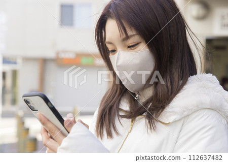 一名戴著口罩的年輕女子在車站前檢查她的智慧型手機 一名戴著口罩的年輕女子在車站前檢查她的智慧型手機 112637482