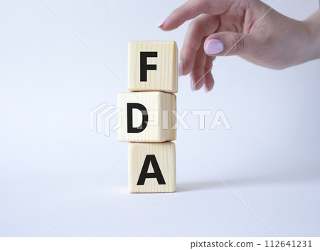 FDA - Food Drug Administration symbol. Wooden cubes with word FDA. Doctor hand. Beautiful white background. Medical and Food Drug Administration concept. Copy space. FDA - Food Drug Administration symbol. Wooden cubes with word FDA. Doctor hand. Beautiful white background. Medical and Food Drug Administration concept. Copy space. 112641231