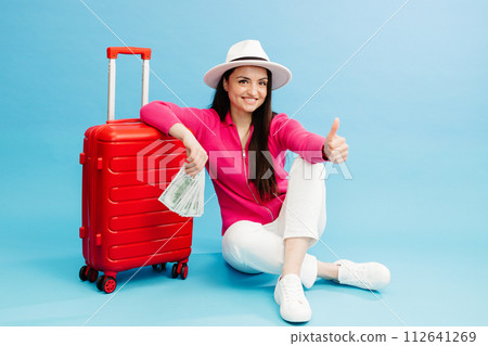 Traveler tourist woman in casual clothes and white hat holding money and sitting at the red suitcase isolated on blue background. Female passenger traveling abroad to travel weekends getaway Traveler tourist woman in casual clothes and white hat holding money and sitting at the red suitcase isolated on blue background. Female passenger traveling abroad to travel weekends getaway 112641269