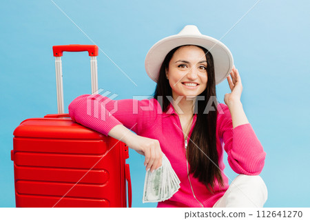 Traveler tourist woman in casual clothes and white hat holding money and sitting at the red suitcase isolated on blue background. Female passenger traveling abroad to travel weekends getaway Traveler tourist woman in casual clothes and white hat holding money and sitting at the red suitcase isolated on blue background. Female passenger traveling abroad to travel weekends getaway 112641270