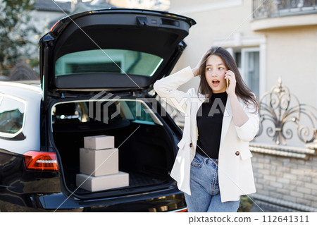 Young business woman standing at the car with parcels, coming home by car. The girl is standing and talking on mobile phone near the car. Concept of buying goods online and delivering them home Young business woman standing at the car with parcels, coming home by car. The girl is standing and talking on mobile phone near the car. Concept of buying goods online and delivering them home 112641311
