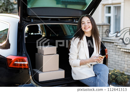 Young business woman sitting in the car, coming home by car. The girl is sitting in the car and holding a mobile phone. Concept of buying goods online and delivering them home Young business woman sitting in the car, coming home by car. The girl is sitting in the car and holding a mobile phone. Concept of buying goods online and delivering them home 112641313