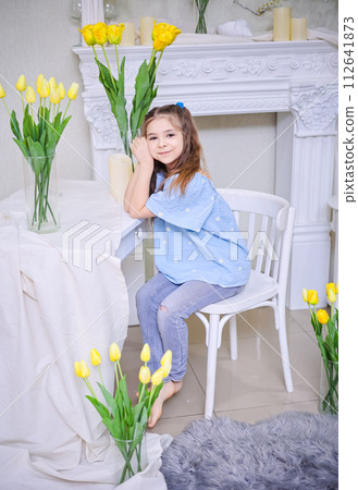 A beautiful girl in a blue blouse sits against the background of bouquets with yellow tulips. A beautiful girl in a blue blouse sits against the background of bouquets with yellow tulips. 112641873
