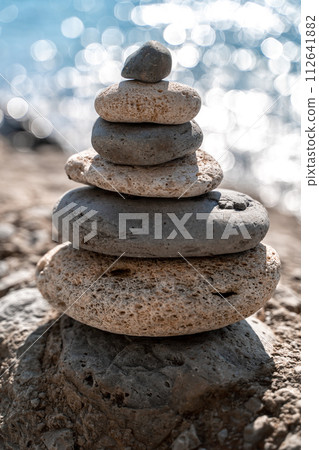 A tower of stones. Balanced pyramid of pebbles on the beach on a sunny day. Blue sea in the background. Selective focus, bokeh. Zen stones on the sea beach, meditation, spa, harmony, tranquility 112641882