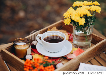A cup of tea or coffee on a wooden tray against the autumn background with fallen leaves, candle and flowers, autumn mood 112643299