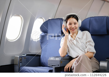 Asian woman sitting and using a smartphone in an airplane Listening to music next to the airplane seat window travel and technology concept Asian woman sitting and using a smartphone in an airplane Listening to music next to the airplane seat window travel and technology concept 112644226
