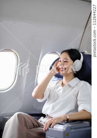 Asian woman sitting and using a smartphone in an airplane Listening to music next to the airplane seat window travel and technology concept Asian woman sitting and using a smartphone in an airplane Listening to music next to the airplane seat window travel and technology concept 112644227