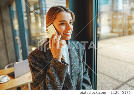 Stylish young female freelancer is talking phone with client while standing in coworking near window 112645175