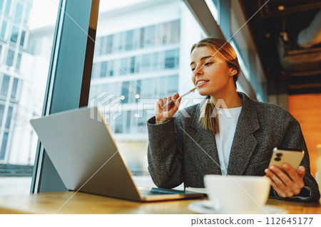 Smiling woman manager working on laptop and holding phone while sitting in cozy cafe 112645177